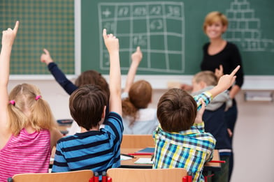 Intelligent group of young school children all raising their hands in the air to answer a question posed by the female teacher, view from behind-1